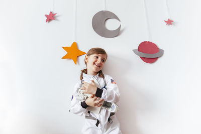Smiling girl wearing spacesuit holding model airplane against wall at home
