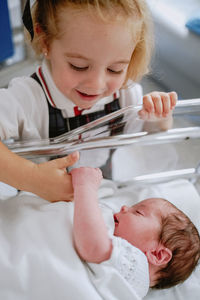 From above of cheerful curious little girl standing near newborn baby sibling sleeping in hospital cradle in ward