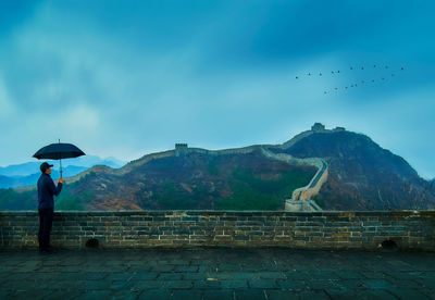 Man with umbrella standing at great wall of china against cloudy sky