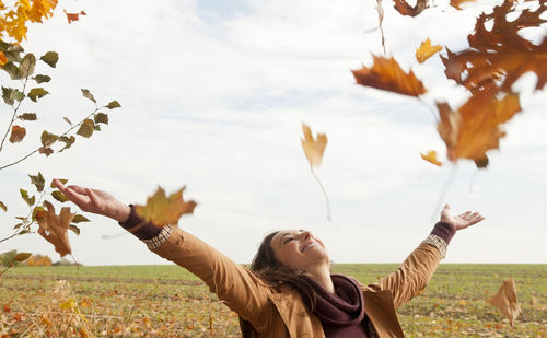 Cheerful woman throwing autumn leaves at park against cloudy sky