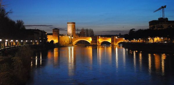 Bridge over river by illuminated buildings against sky at dusk