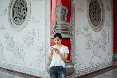 Smiling young woman using smart phone standing against wall