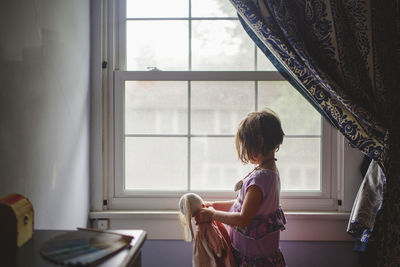 A small child stands in window light holding soft toy bunny