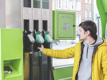 Beautiful young caucasian man in a yellow jacket and gray sports take a filling gun at a gas station