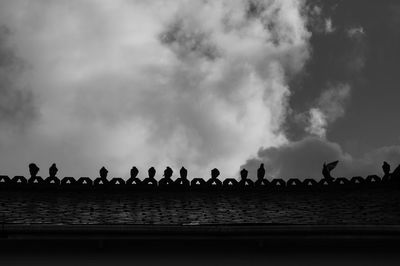Silhouette birds perching on boardwalk against cloudy sky