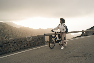 Young woman wheeling cycle on road against sky