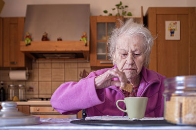 Very old woman, alone in her kitchen, dips cookie in cup of latte coffee, during breakfast