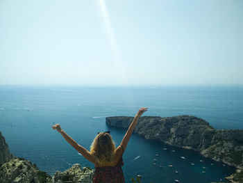 Rear view of woman with arms raised sitting on cliff by sea against sky