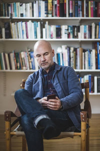 Confident mature therapist using mobile phone while sitting against bookshelf at home office