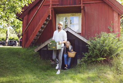 Full length of friends holding vegetable crate while standing against cottage