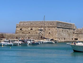 Sailboats moored on sea by buildings against clear blue sky