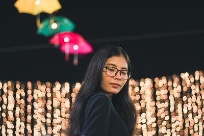 Portrait of young woman standing by illuminated lighting equipment at night