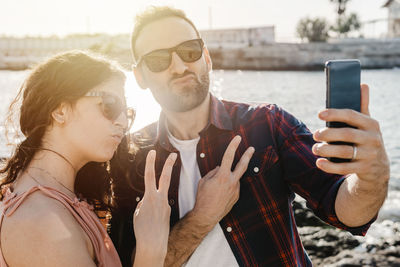 Couple wearing sunglasses taking selfie with smart phone against lake at sunset
