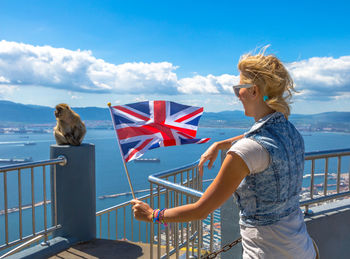 Woman holding flag while standing by railing against sky