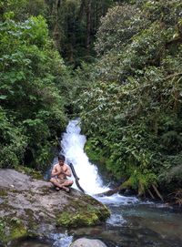 Shirtless man meditating on rock against stream in forest