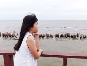 Girl standing at railing by beach against sky