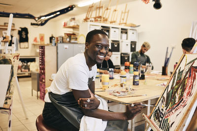Portrait of smiling young male student painting on artist's canvas in art class