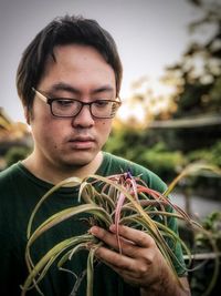 Portrait of young man holding flowering air plant against sky and trees.