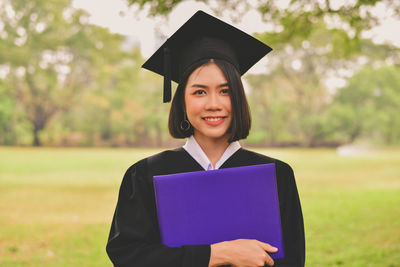 Portrait of young woman in graduation gown holding file while standing at park