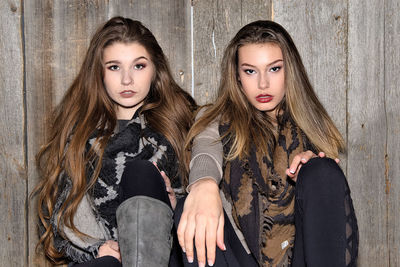 Portrait of sisters wearing warm clothing while sitting against wooden wall