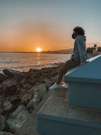 Man sitting on retaining wall against sea during sunset