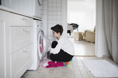 Side view of boy loading washing machine while crouching at home