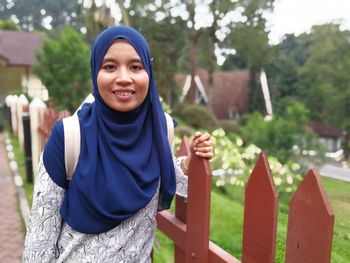 Portrait of smiling mid adult woman standing by fence against trees in park