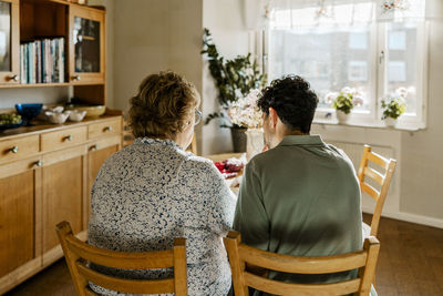 Rear view of young male nurse sitting next to elderly woman on chair in kitchen at home