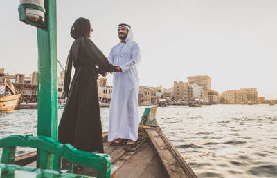 Cheerful couple holding hands while standing in boat on river at sunset