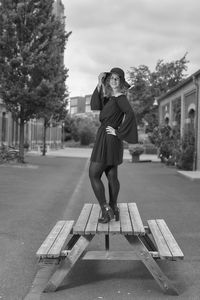 Portrait of young woman standing on bench against sky