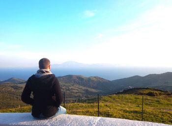 Rear view of man sitting on retaining wall against clear sky