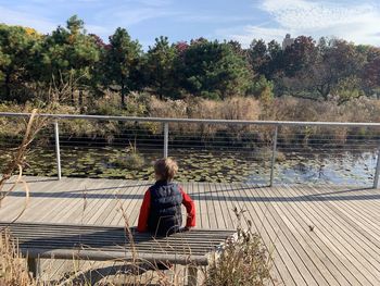 Low angle view of boy sitting on bench looking at a pond in park setting during sunny winter day