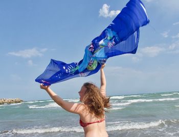 Rear view of teenage girl holding scarf while standing at beach against blue sky