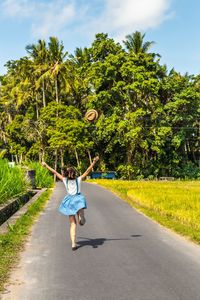 Rear view of woman catching hat while running on road against trees