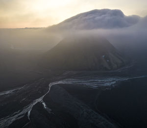 Volcanic mountain with steep surface covered with fog located near flowing river in wild nature against cloudless sky in evening time