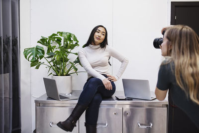 Female blogger photographing colleague sitting on sideboard in creative office