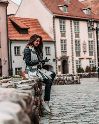 Young woman sitting on stone wall against buildings