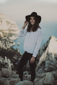 Portrait of smiling young woman wearing hat standing on rocks against sky