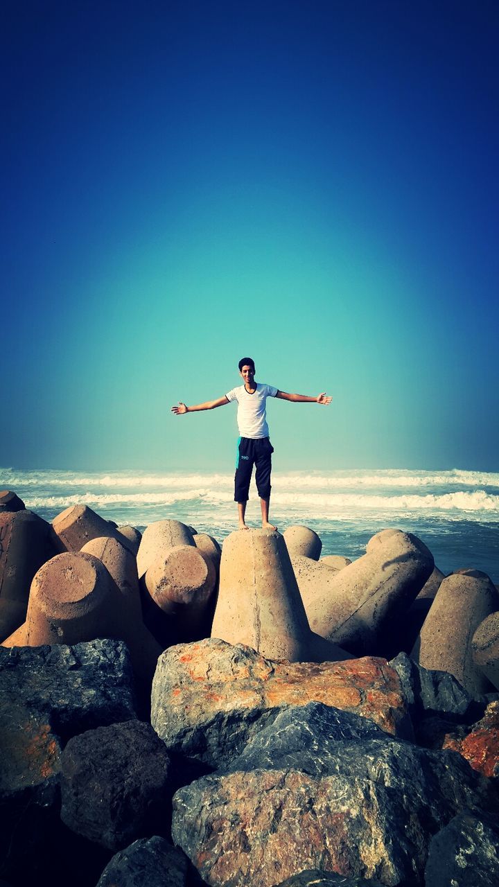 full length, sea, clear sky, lifestyles, leisure activity, copy space, water, standing, rock - object, horizon over water, rear view, casual clothing, tranquility, tranquil scene, scenics, beauty in nature, rock, shore