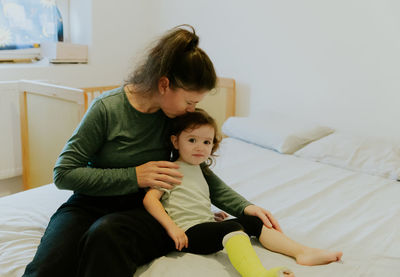 A young girl kisses a child with a cast on her leg sitting on the bed.