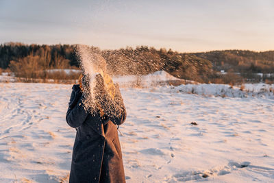 A woman with a obscured face throws snow with her hands in a snow-covered field