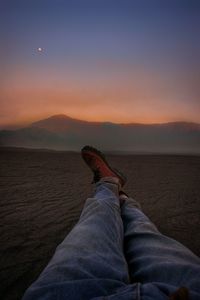 Low section of person relaxing on shore against sky during sunset