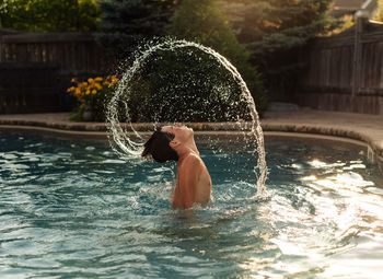 Teenage boy flipping water from his hair in an arc in a backyard pool.