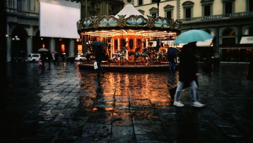 Illuminated carousel against buildings during rainy season