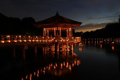 Illuminated building by lake against sky at dusk