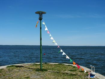 Bunting flags hanging amidst bollard and lamp post by sea against blue sky