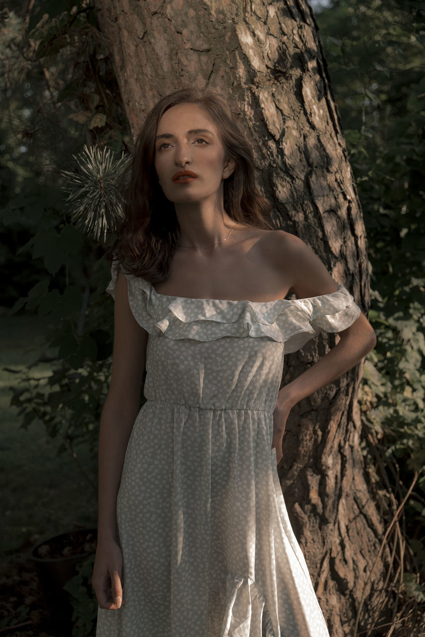 women, fashion, one person, dress, adult, tree, clothing, young adult, wedding dress, portrait, hairstyle, gown, plant, long hair, bridal clothing, standing, photo shoot, nature, forest, female, looking at camera, three quarter length, spring, front view, elegance, brown hair, looking, land, contemplation, outdoors, tree trunk, bride, glamour, serious, trunk, strapless, blond hair, cocktail dress