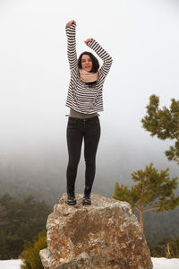 Full length of carefree young woman with arms raised standing on rock during foggy weather