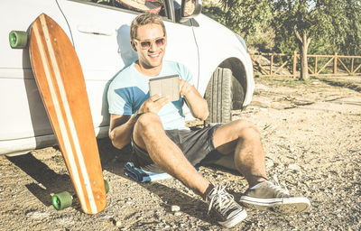 Young man using digital tablet while sitting by skateboard against car