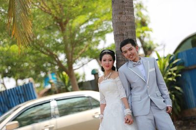 Portrait of smiling bride and groom standing against tree trunk